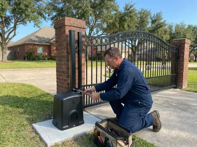 Gate mechanic kneeling beside a gate opener at a driveway swing gate, tightening the arm bracket with a wrench