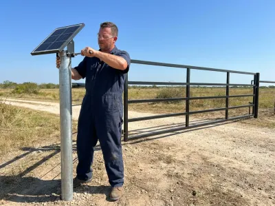 Gate mechanic tightening the bracket of a small solar panel on a steel post with a wrench, beside a ranch gate